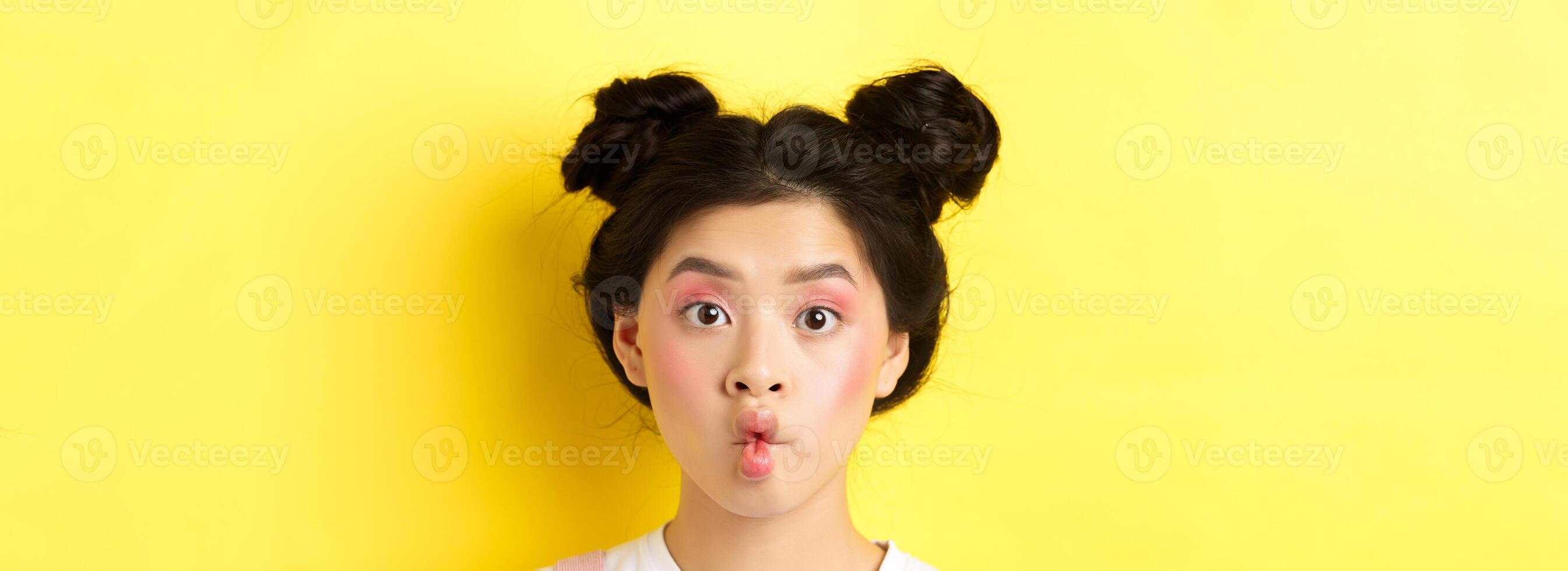 Close-up of teen korean girl pucker lips and looking funny at camera, standing with glamour makeup and stylish hairstyle, yellow background photo