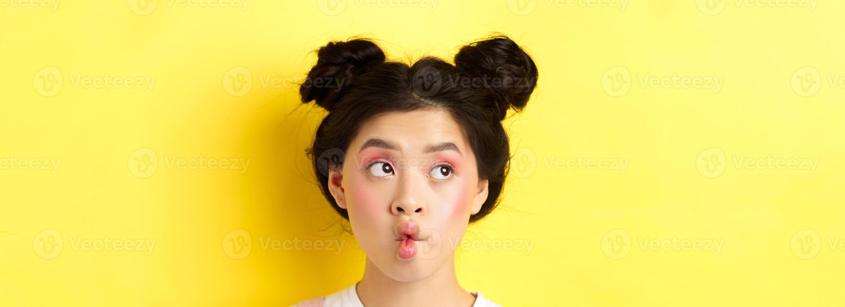 Close-up of teen asian girl pucker lips and looking funny at camera, standing with glamour makeup and stylish hairstyle, yellow background photo