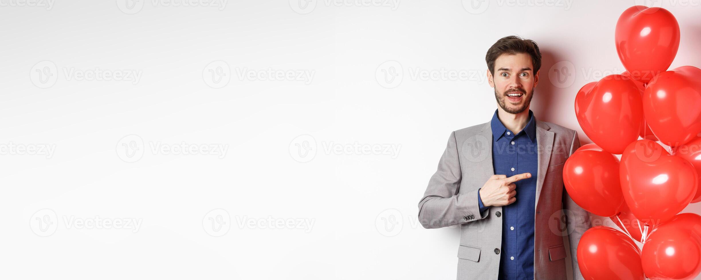 Lovers day. Excited male model in suit pointing finger at Valentines heart balloons and smiling, prepare romantic gifts on date, standing over white background photo