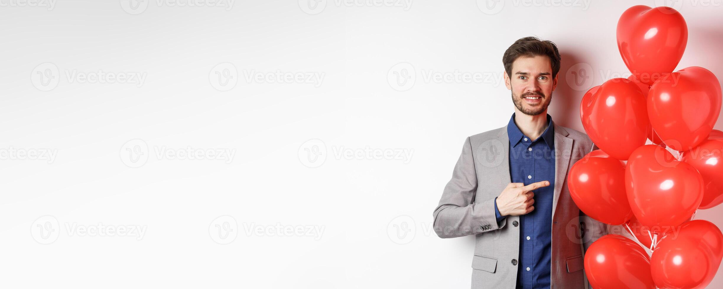 Lovers day. Handsome smiling guy in suit making surprise gift on Valentines date, pointing at romantic heart balloons and looking happy, standing over white background photo