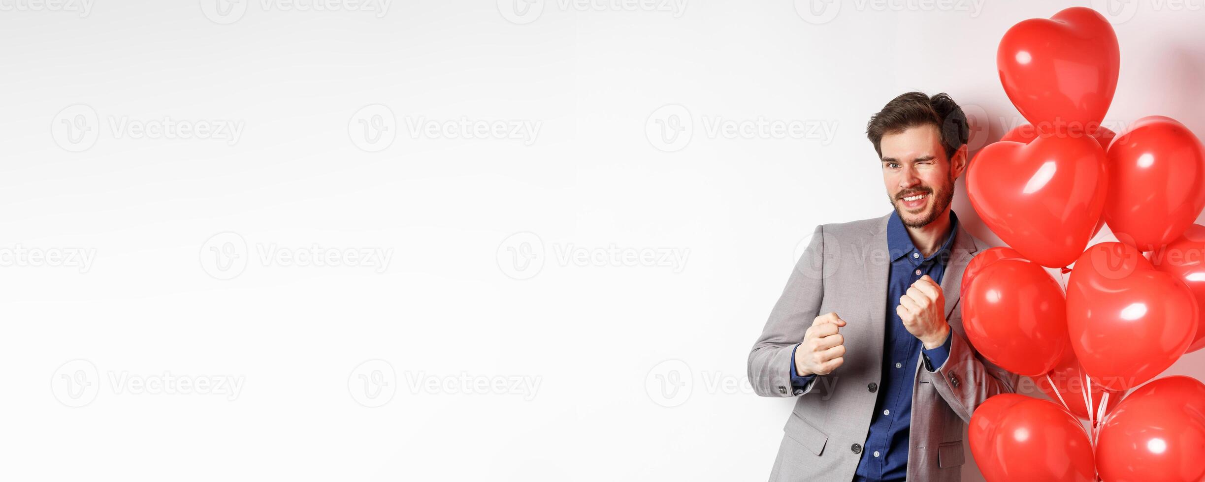 Handsome man dancing and smiling near Valentines day hearts balloon, winking at camera, getting dressed on romantic date on lovers holiday, white background photo