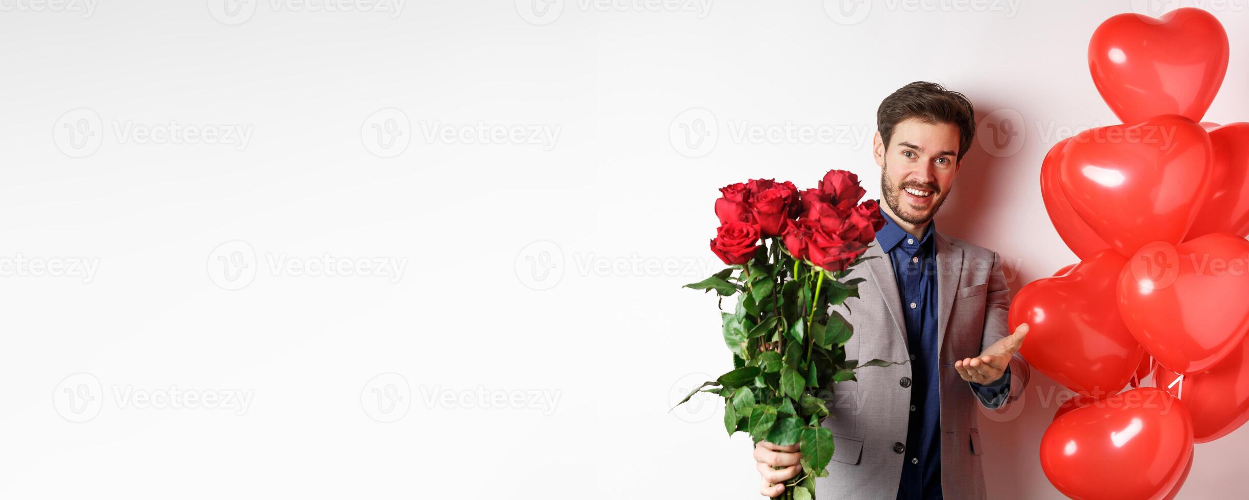 Romantic guy in suit saying happy Valentines day, giving red roses to you and pointing hand, standing with heart balloon gift over white background photo