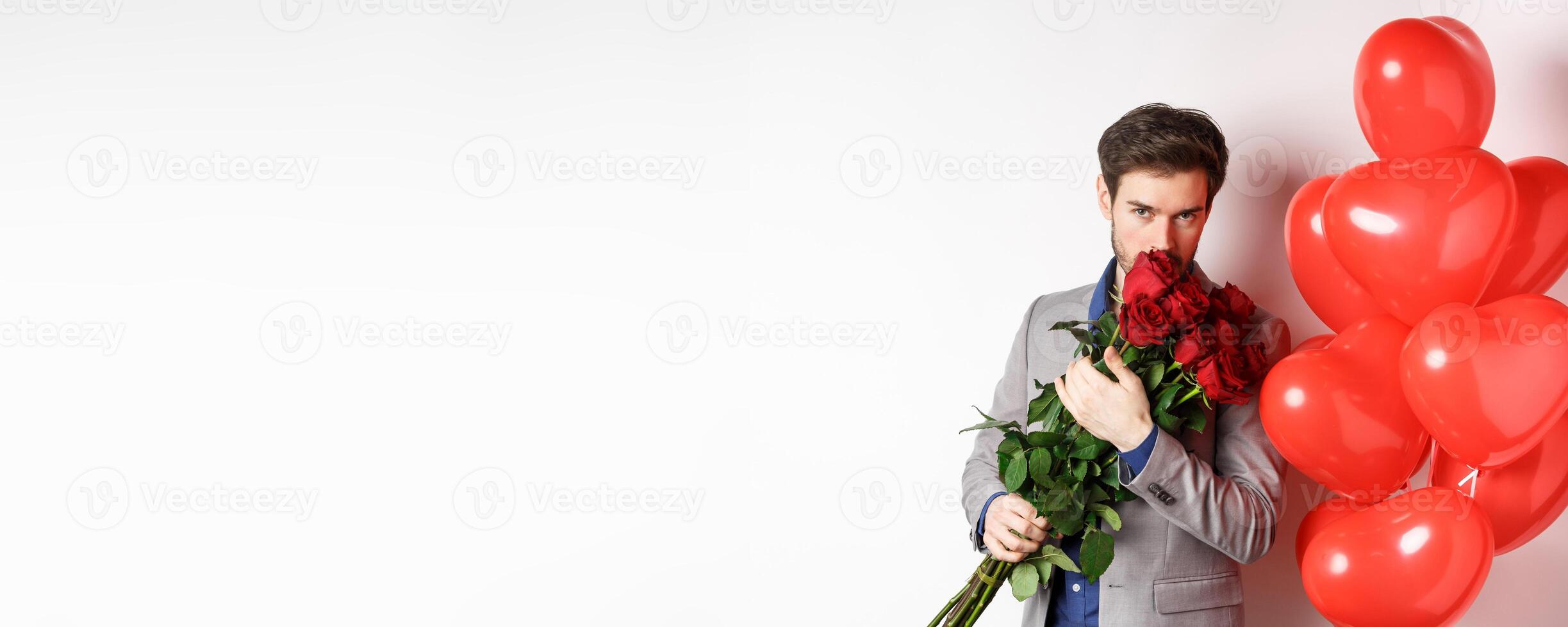 Romantic man smell bouquet of red roses and looking passionate at camera. Boyfriend in suit going on Valentines date with gifts and heart balloons, white background photo
