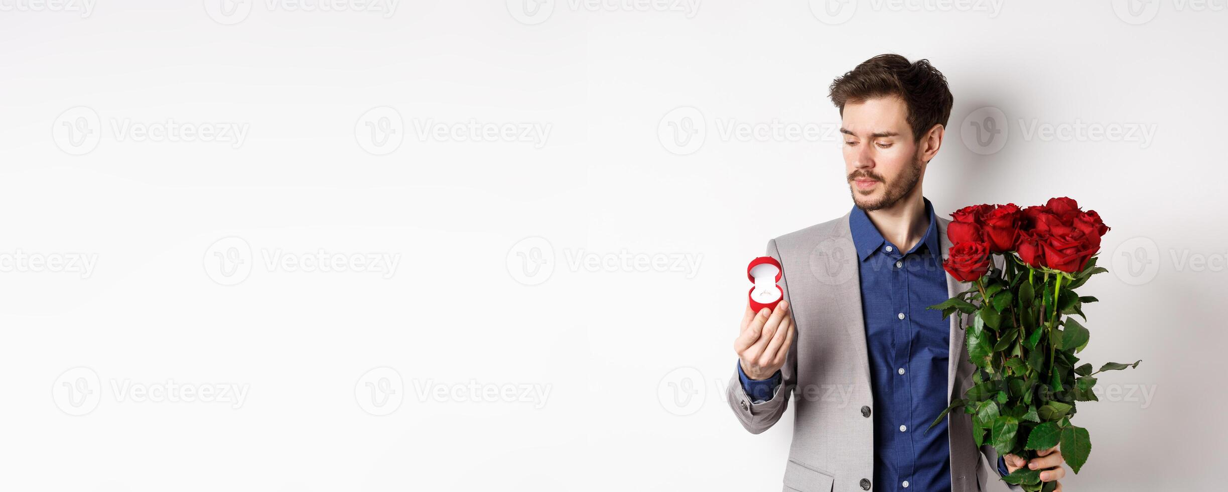 Handsome bearded man in suit looking at engagement ring, making surprise on lovers day, standing with red roses over white background photo