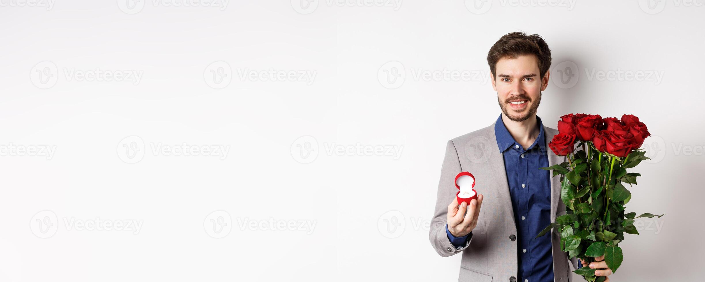 Smiling boyfriend making a marriage proposal, standing with engagement ring and bouquet of red roses, going on romantic Valentines day date, white background photo