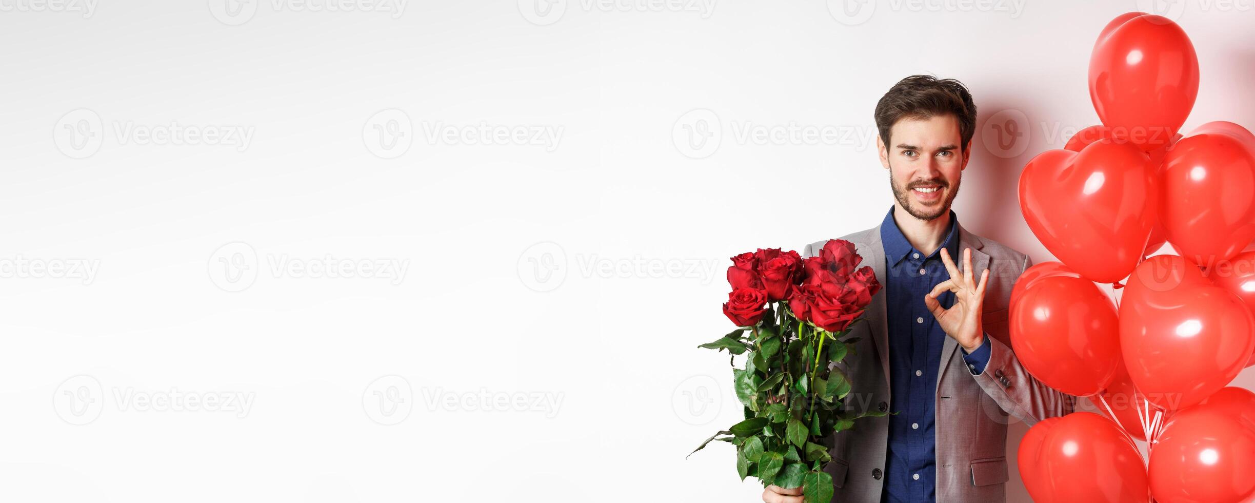 Young man in suit have all under control, showing OK sign and holding red roses, prepare romantic surprise with heart balloons and flowers on Valentines day, white background photo