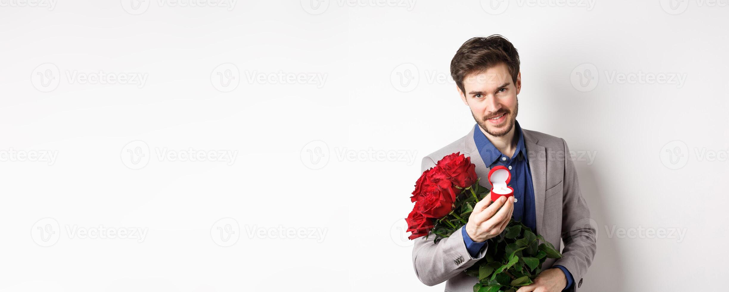 Romantic man with boquet of red roses asking to marry him, holding engagement ring and looking confident at camera, standing in suit over white background photo