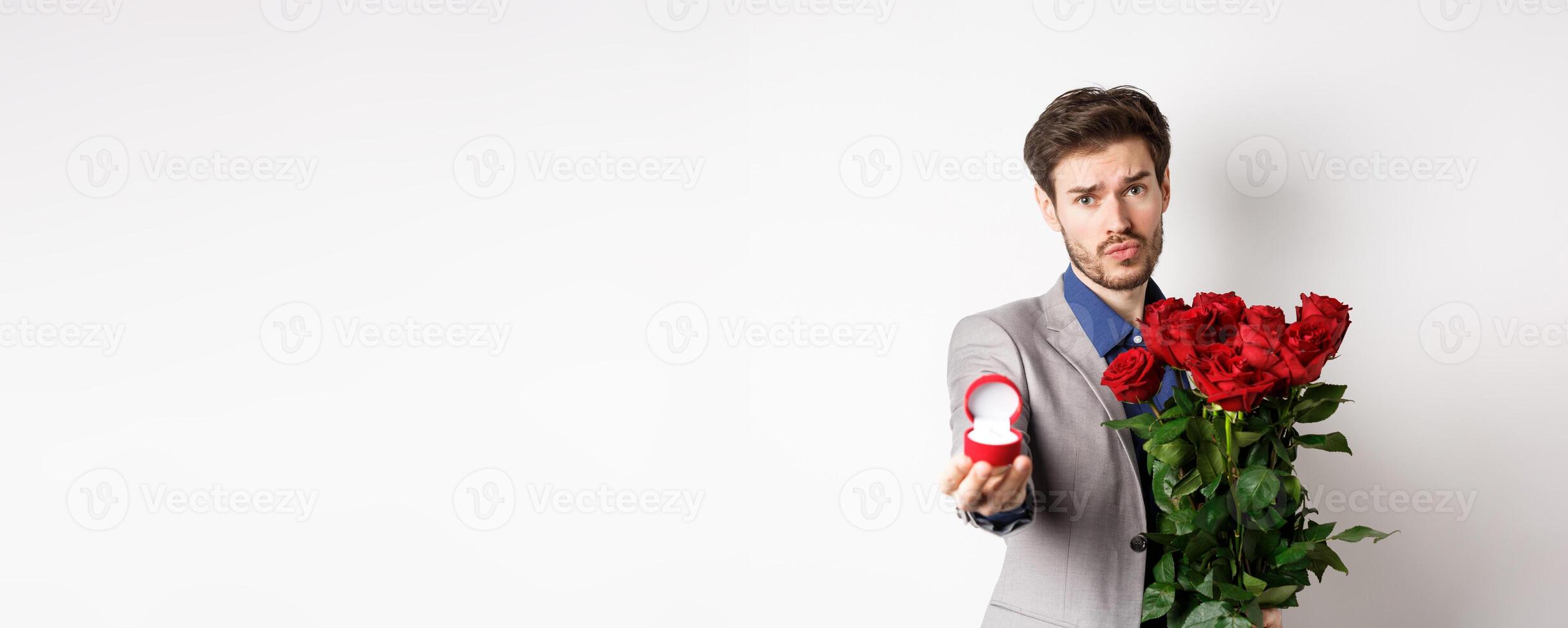 Handsome boyfriend in suit asking to marry him, standing with red bouquet of roses and engagement ring, looking pleading at camera, standing over white background photo