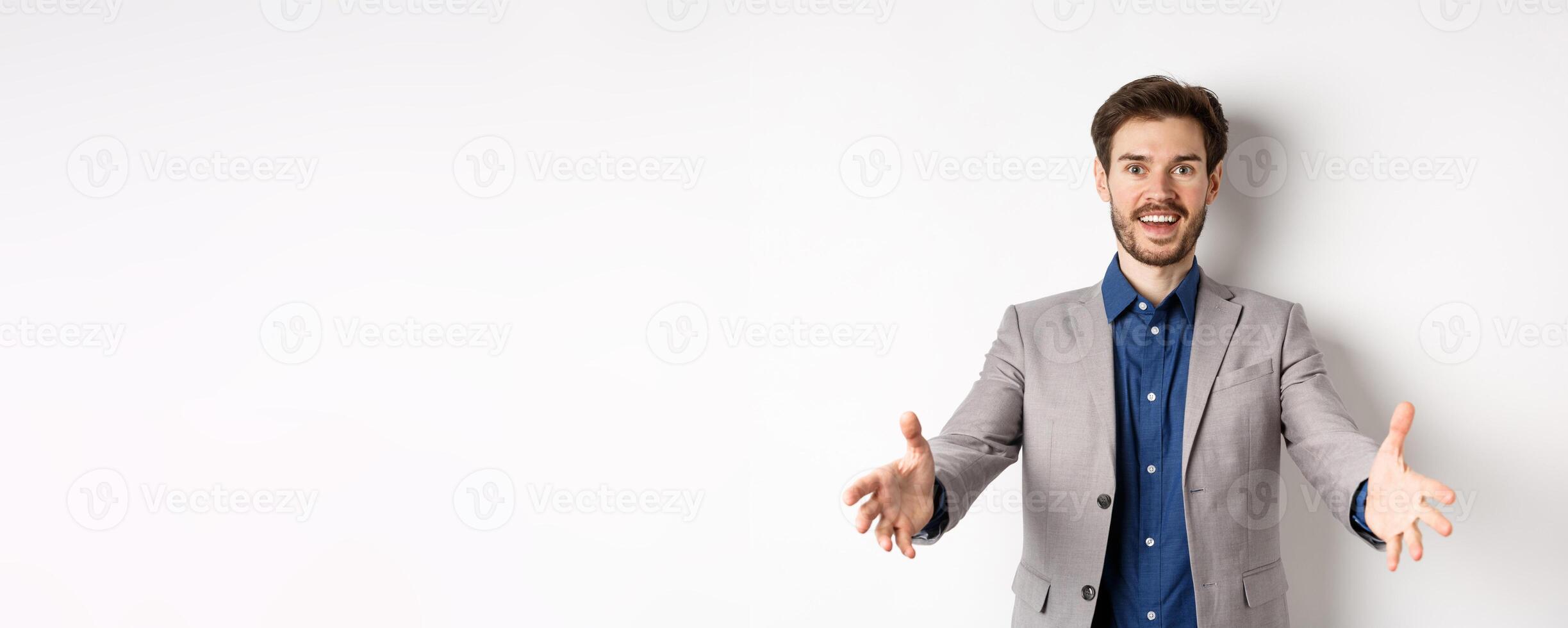 Happy handsome man in suit stretch out hands to greet business partners, welcome guests and smiling excited, standing on white background photo