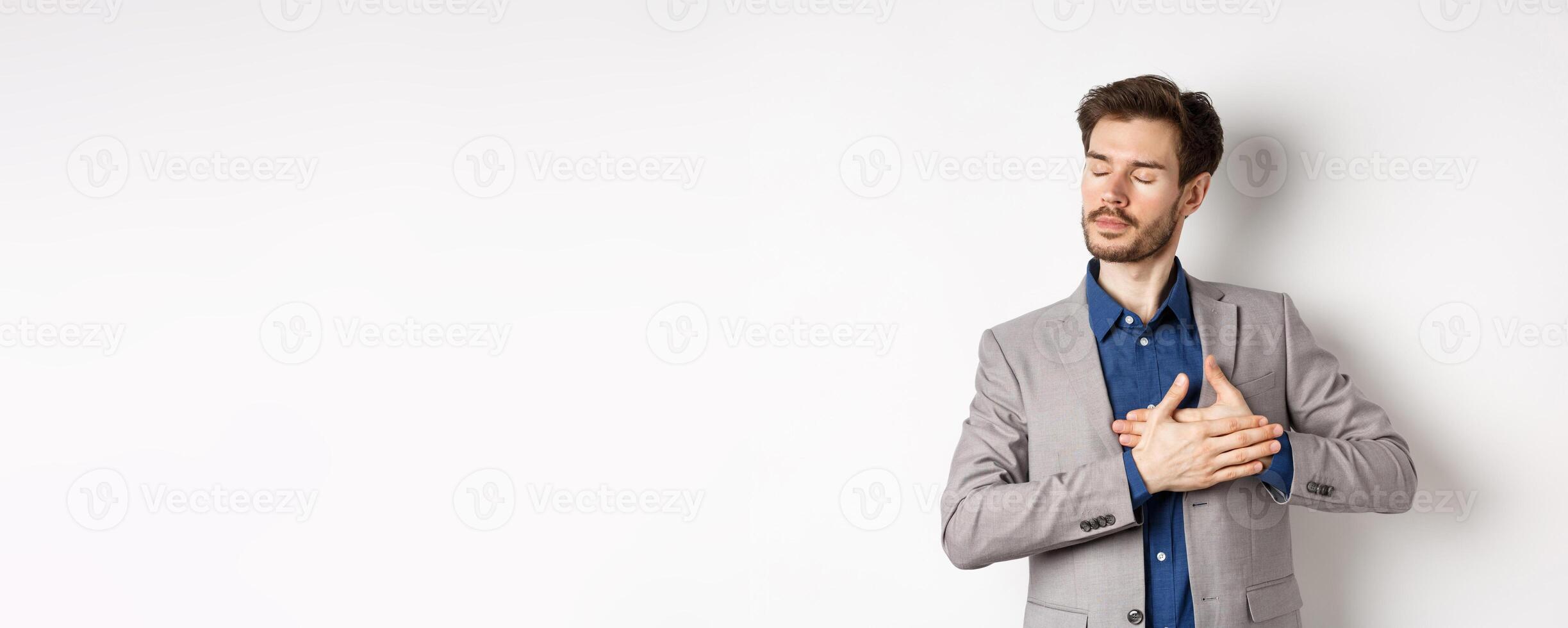 Romantic bearded man in stylish suit holding hands on heart, close eyes and remember something with tender feelings, standing nostalgic on white background photo
