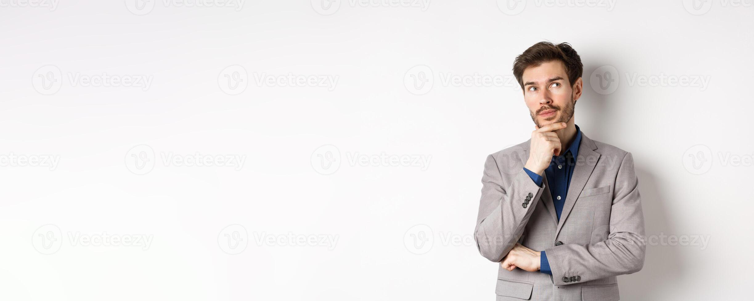 Pensive handsome man in suit looking up and thinking, making choice, pondering ideas, standing against white background photo