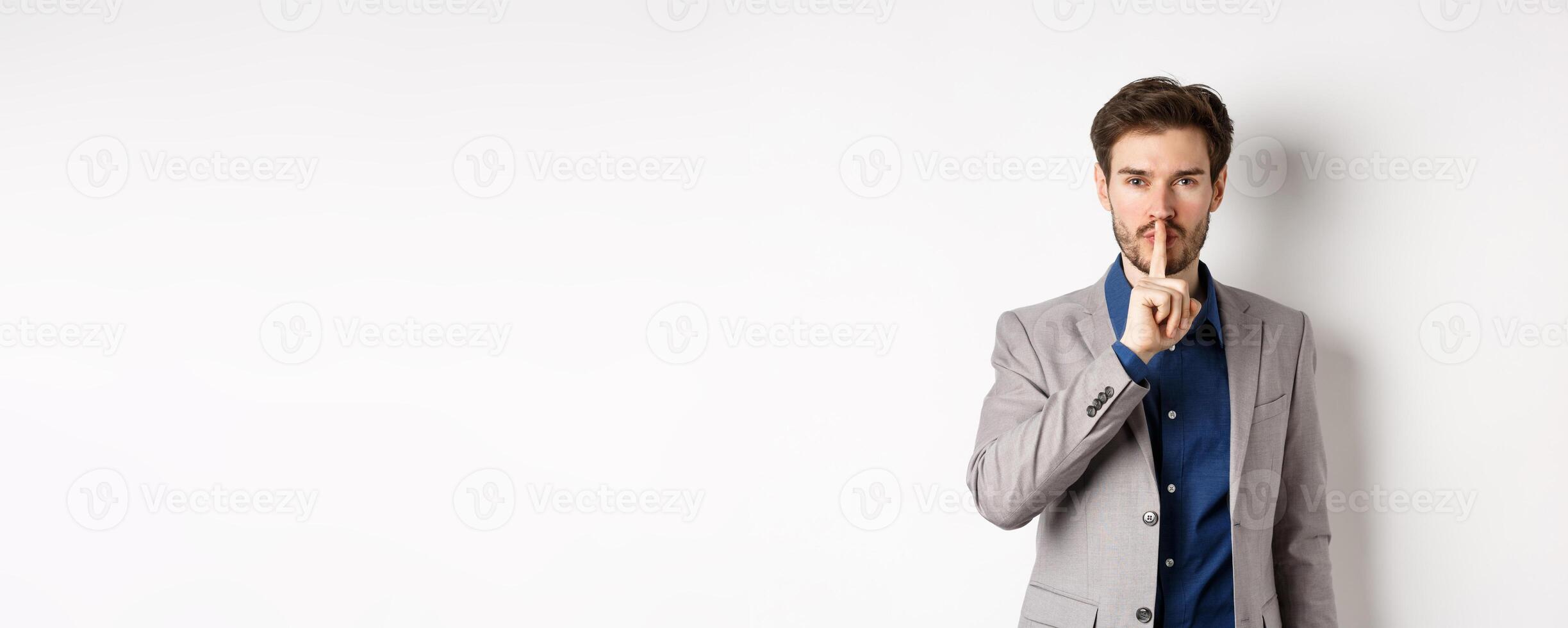 Handsome macho man in suit shushing at camera, press finger to lips, ask keep voice down, tell secret, standing against white background photo