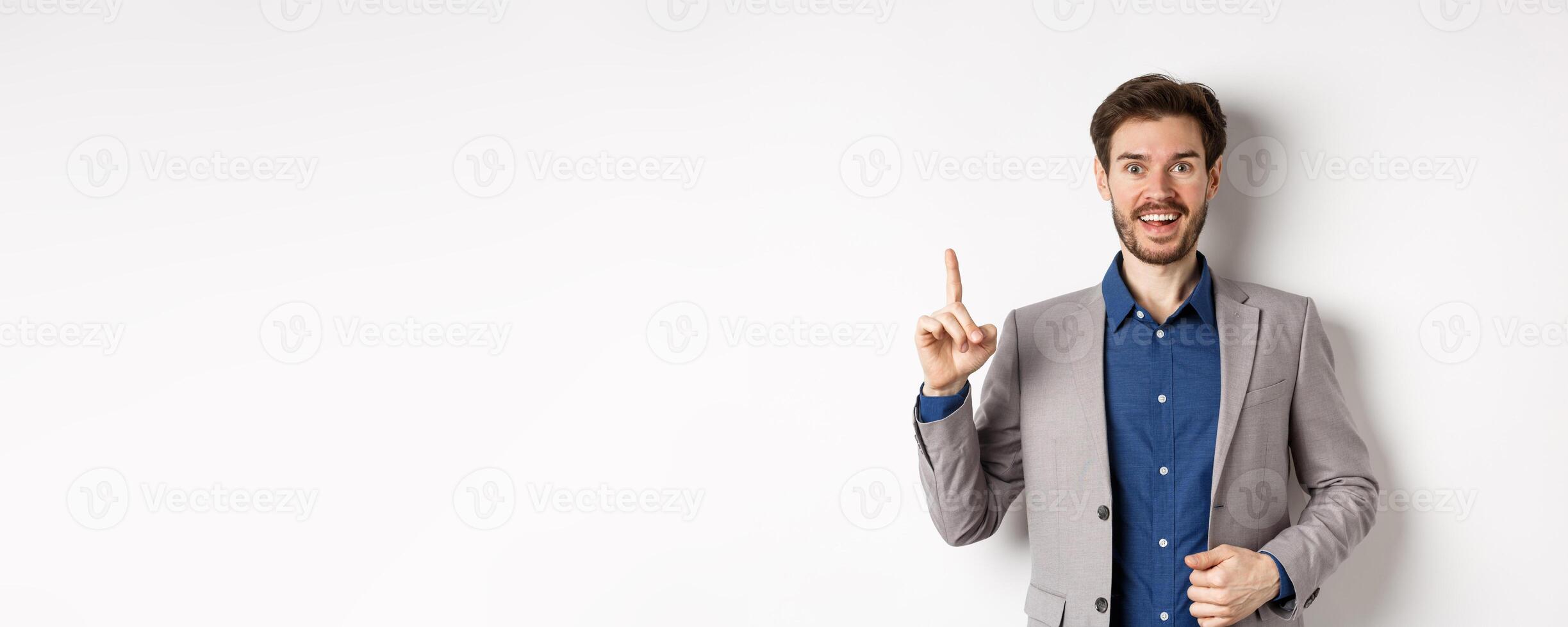 Excited businessman in suit pitching good idea, raising finger in eureka sign and looking amused, saying suggestion, standing on white background photo