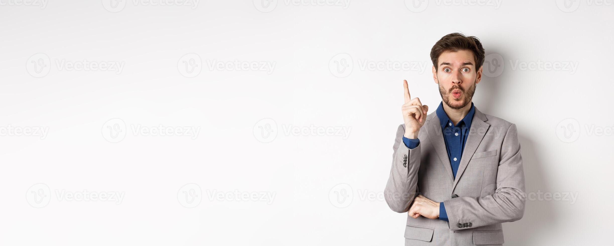Excited businessman in suit pitching good idea, raising finger in eureka sign and looking amused, saying suggestion, standing on white background photo