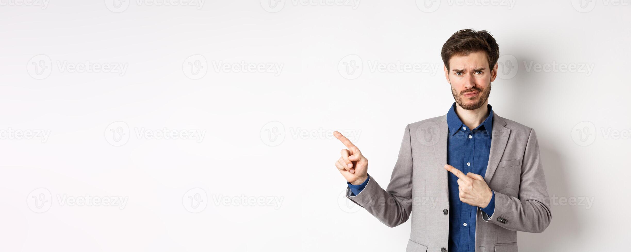 Bad idea. Displeased skeptical guy in business suit smirk and frown, pointing fingers left at upsetting news, looking hesitant at camera, white background photo