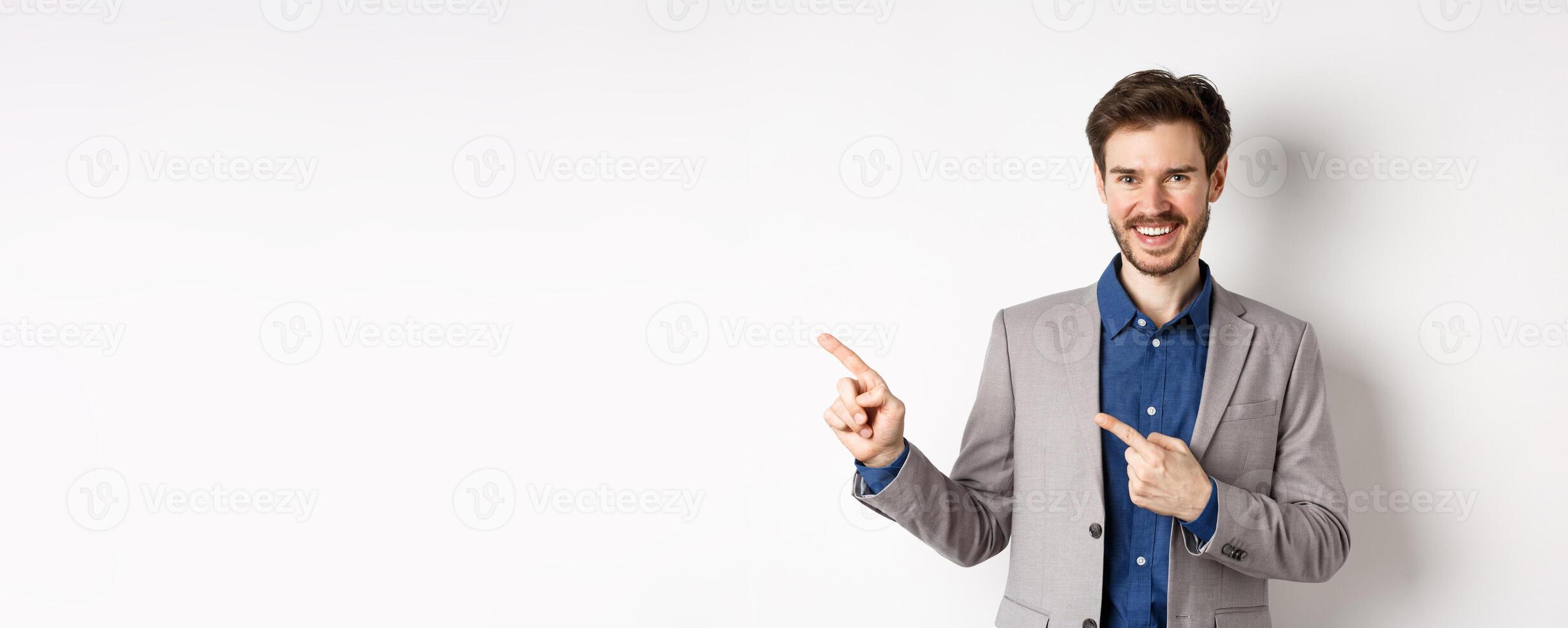 Handsome caucasian man in business suit pointing fingers left at banner and smiling, showing advertisement, standing against white background photo
