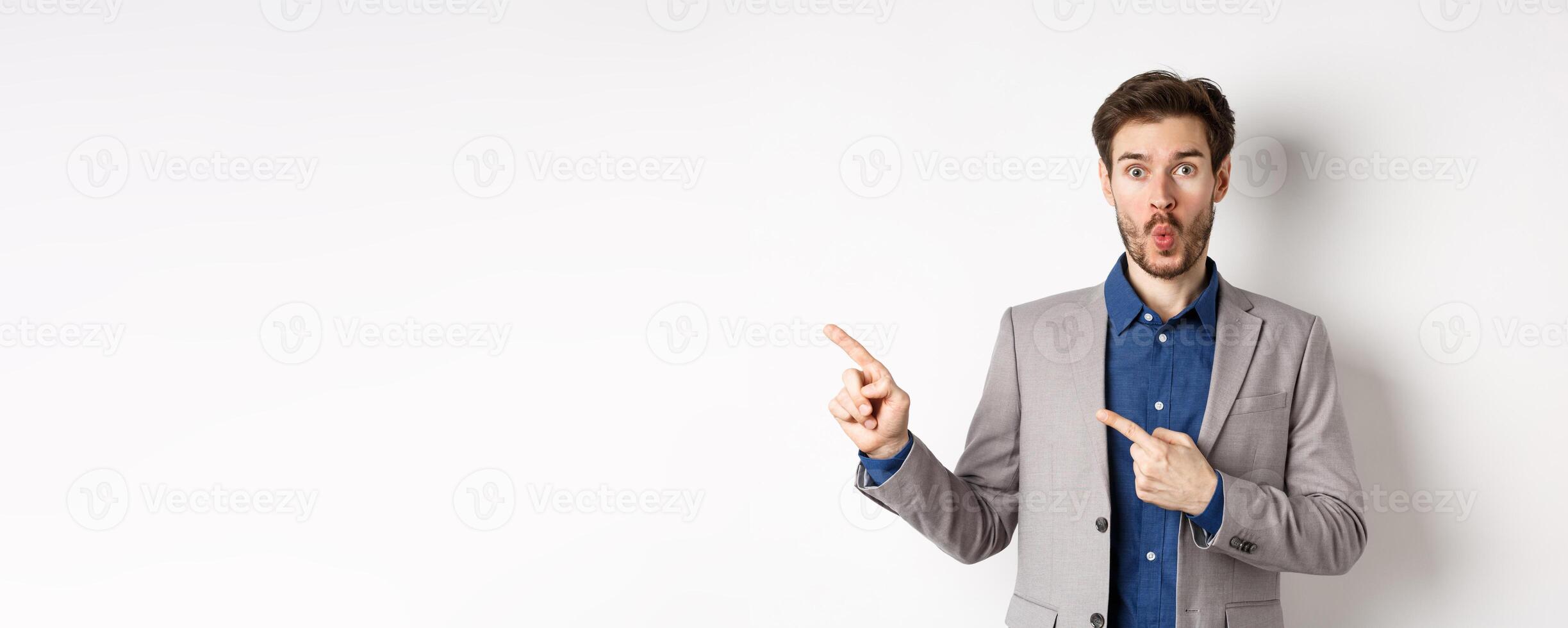Excited handsome man in suit pointing fingers left at banner and saying wow, look here gesture, showing advertisement, standing on white background photo