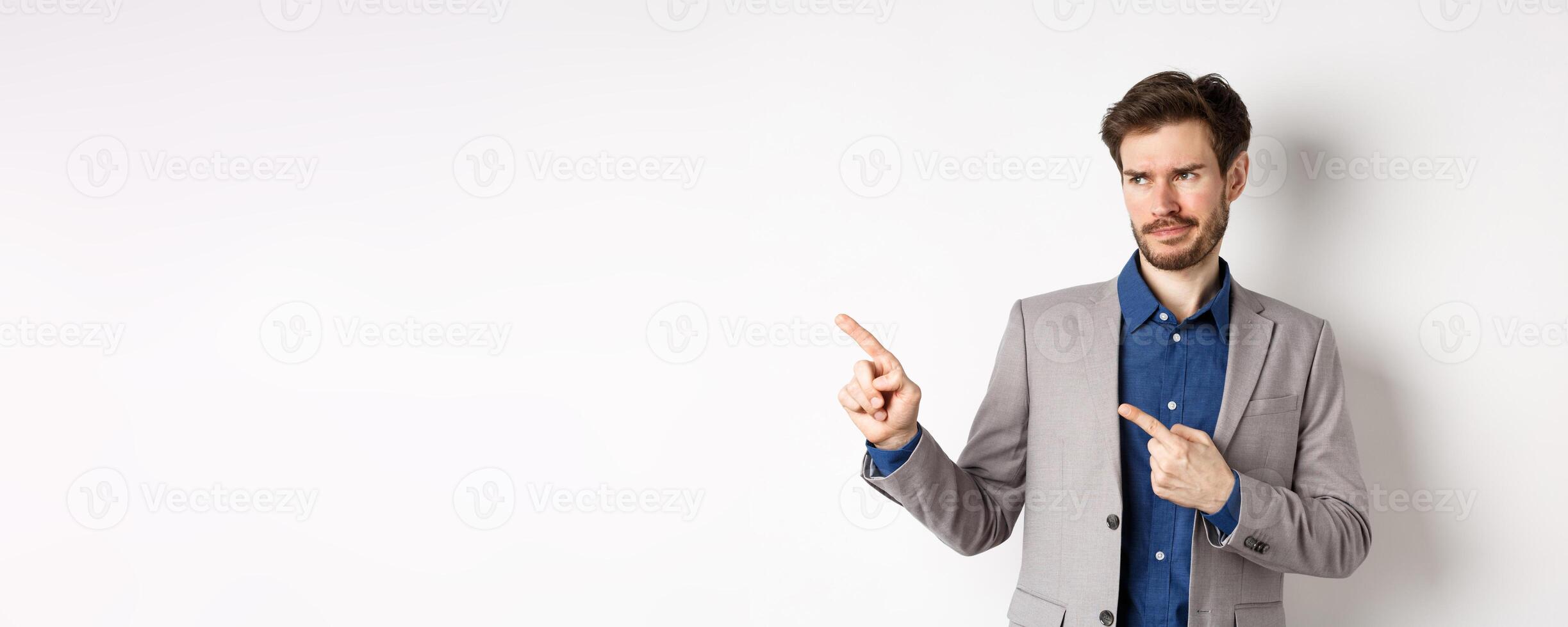Displeased businessman in suit pointing and looking left with skeptical smirk, dislike banner, disapprove promo offer, standing on white background photo