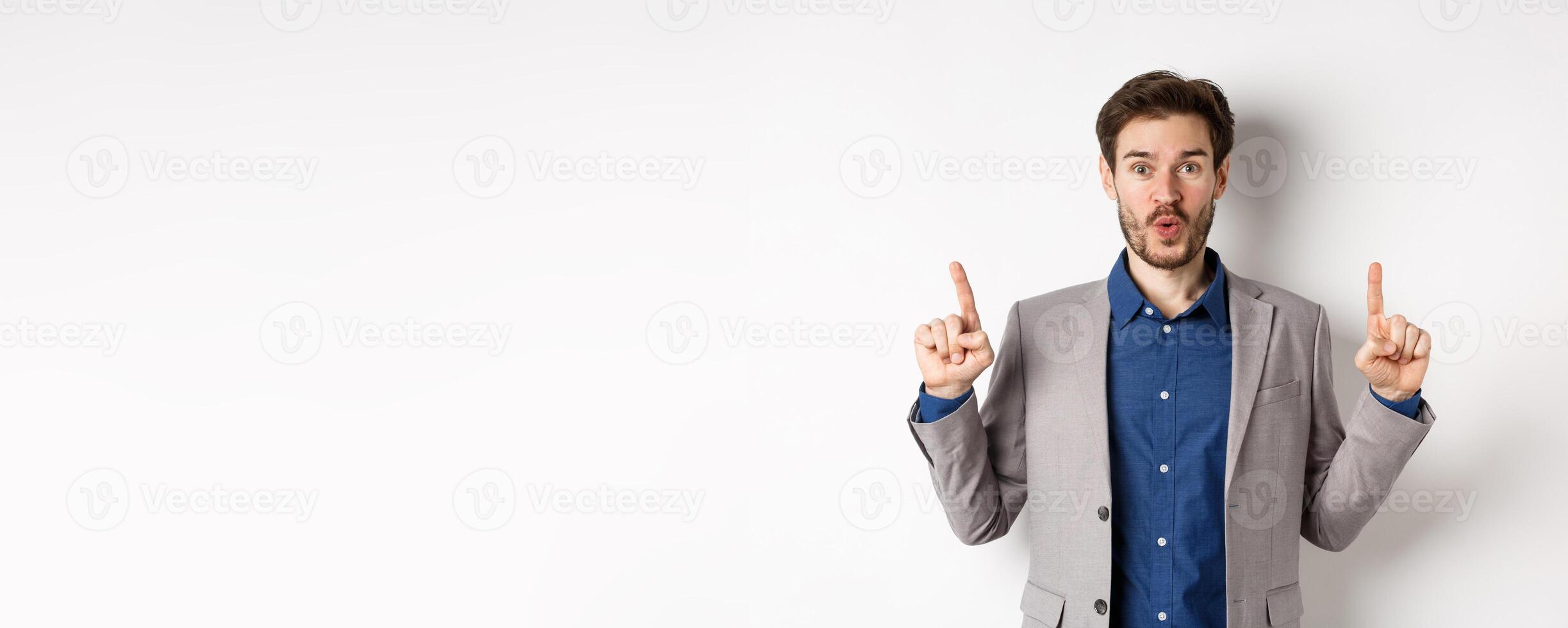 Excited businessman in suit say wow and smiling amused, pointing fingers up at good deal, standing against white background photo