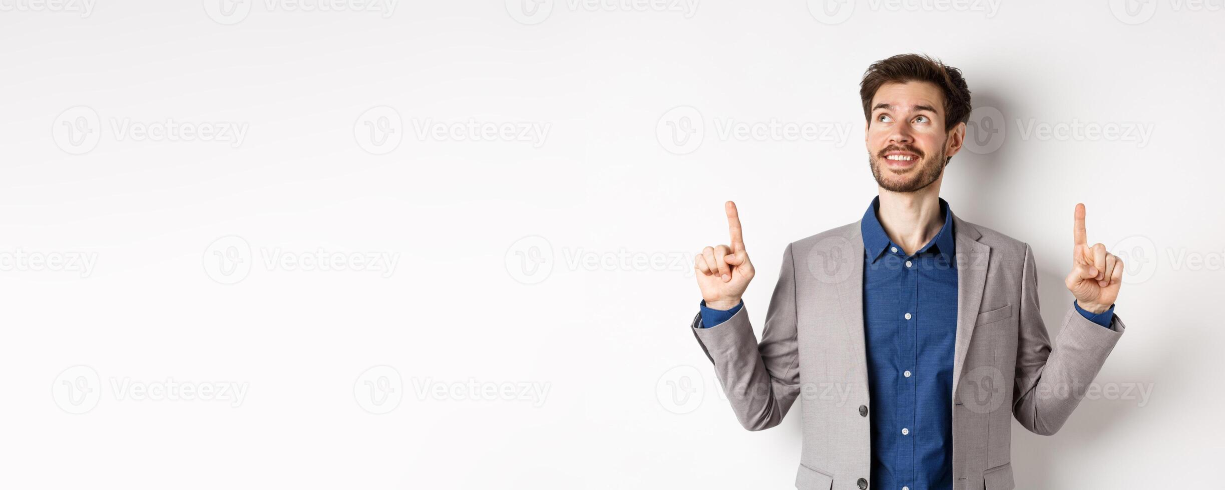 Dreamy smiling male manager in suit pointing and looking up, happy face, checking out advertisement, standing against white background photo