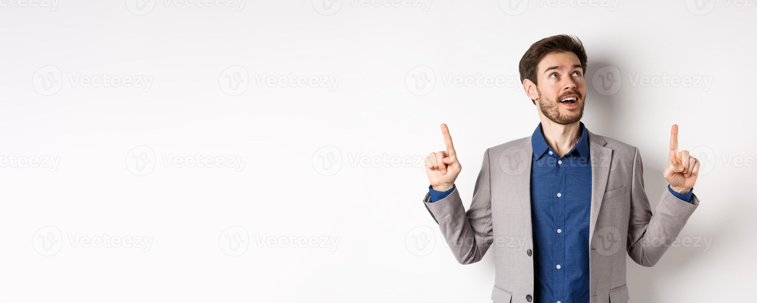 Excited handsome man with beard, wearing suit and looking amused up, pointing at top with admiration, standing against white background photo