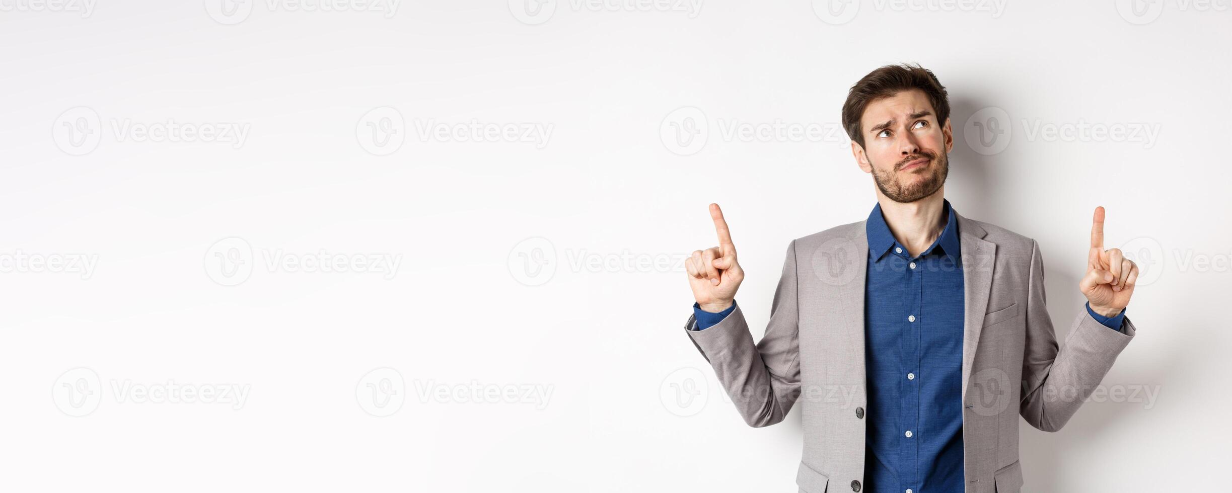 Skeptical and hesitant young businessman in suit looking up with disappointed smirk, feel unsure about deal, standing dissatisfied on white background photo