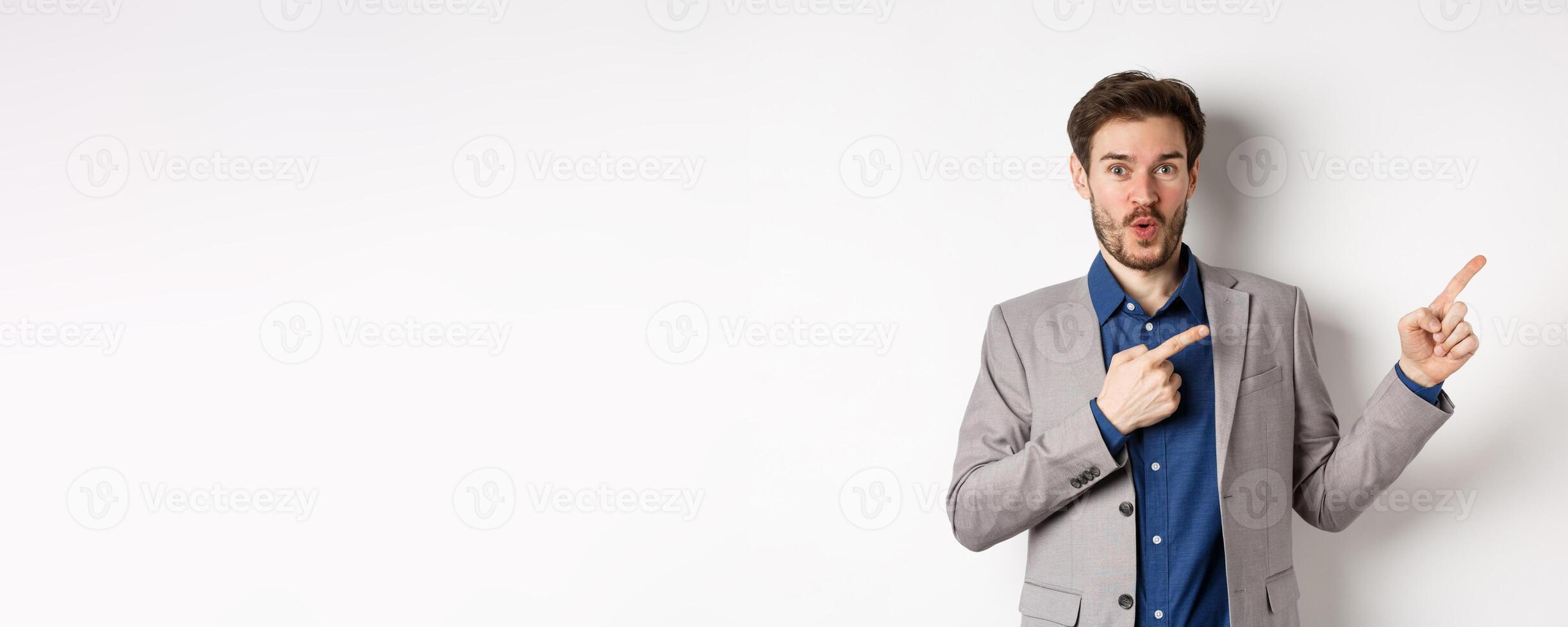 Excited handsome caucasian businessman in suit pointing right at logo, say wow and look amazed, standing on white background photo