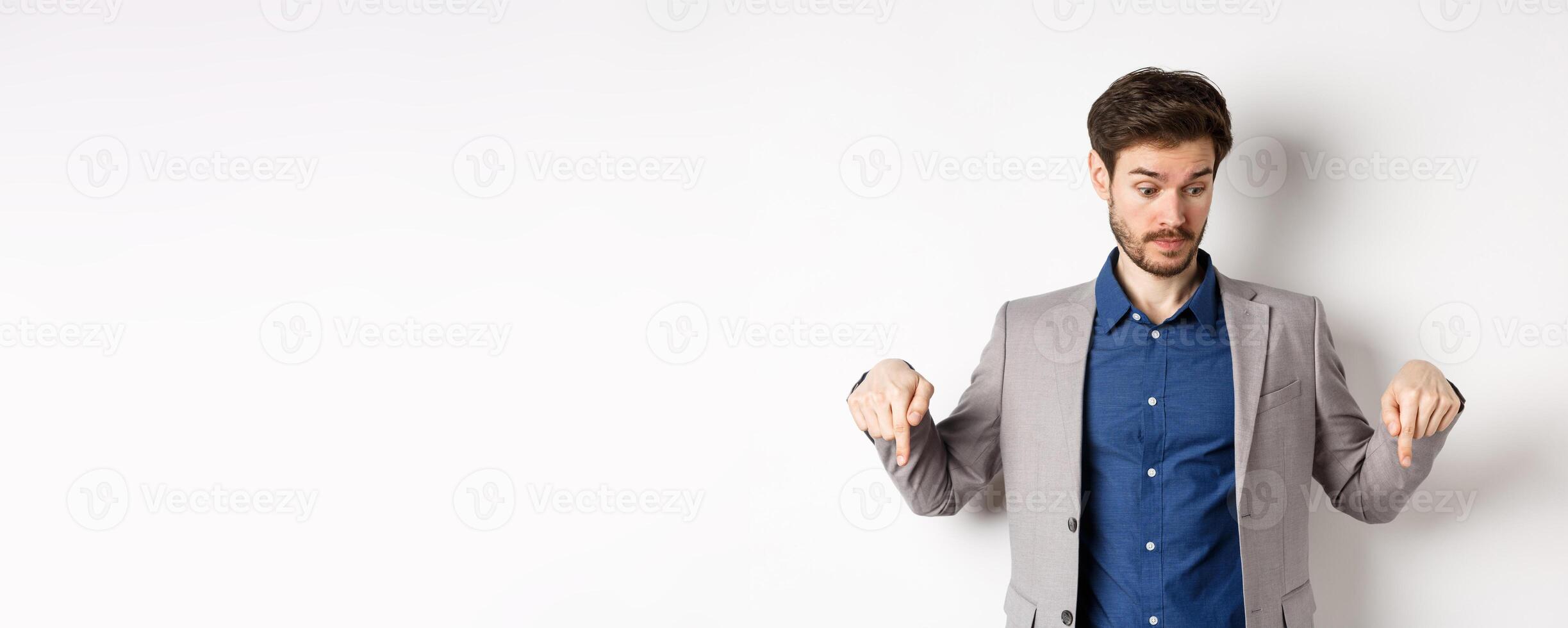 Surprised businessman looking and pointing down with raised eyebrows, see something curious, standing on white background in suit photo