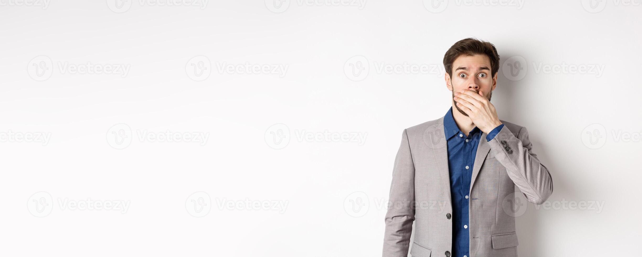 Shocked boyfriend in suit gasping, covering mouth with hand and pop eyes at camera startled, standing on white background photo