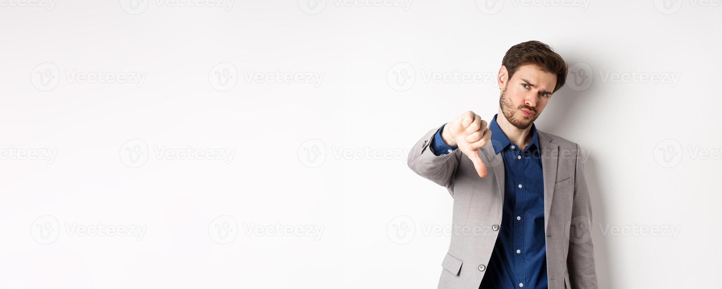Negative emotions. Disappointed businessman frowning, showing thumb down to disapprove product, dislike something bad, stading on white background photo