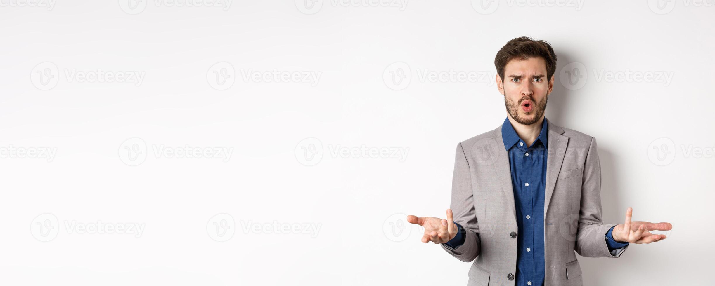 Confused and shocked businessman raising hands up and asking what happened, frowning disappointed, waiting for explanation, standing in suit on white background photo