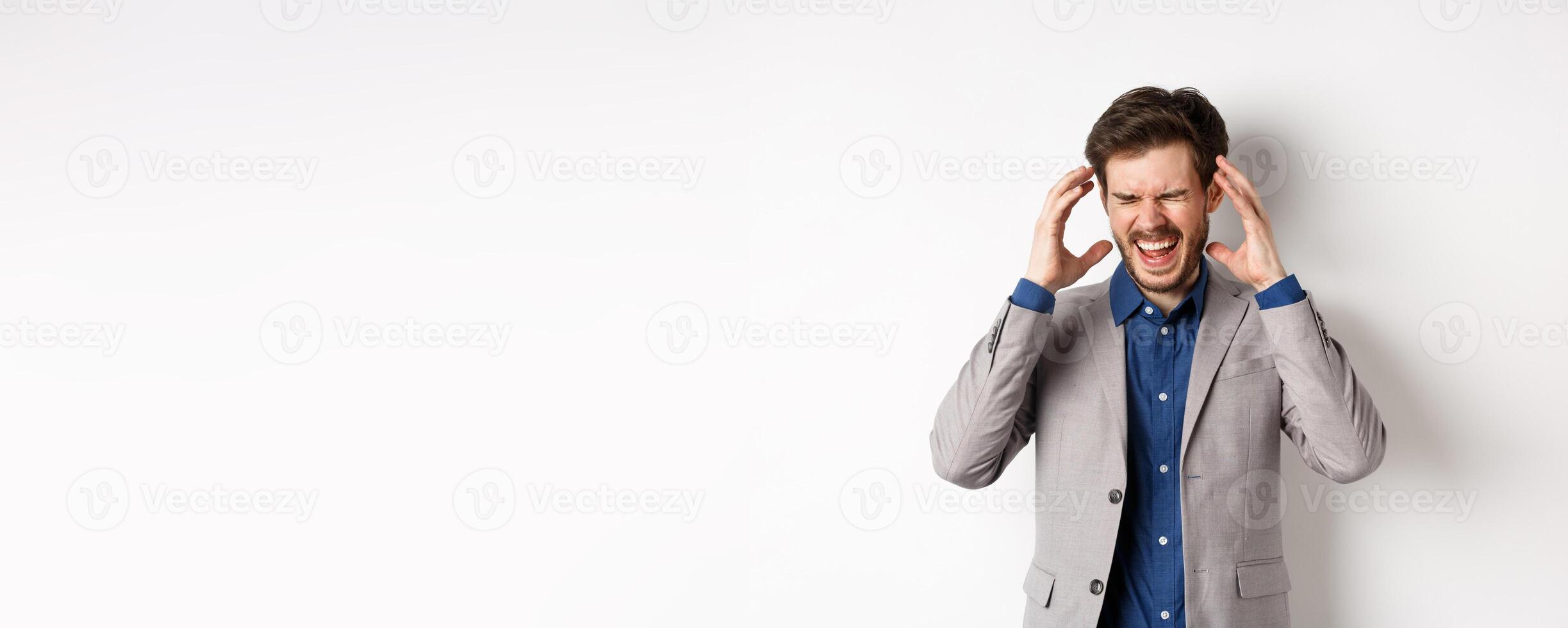 Frustrated business man in suit going crazy, screaming and shaking hands near head, feeling annoyed or angry, shouting distressed, standing on white background photo