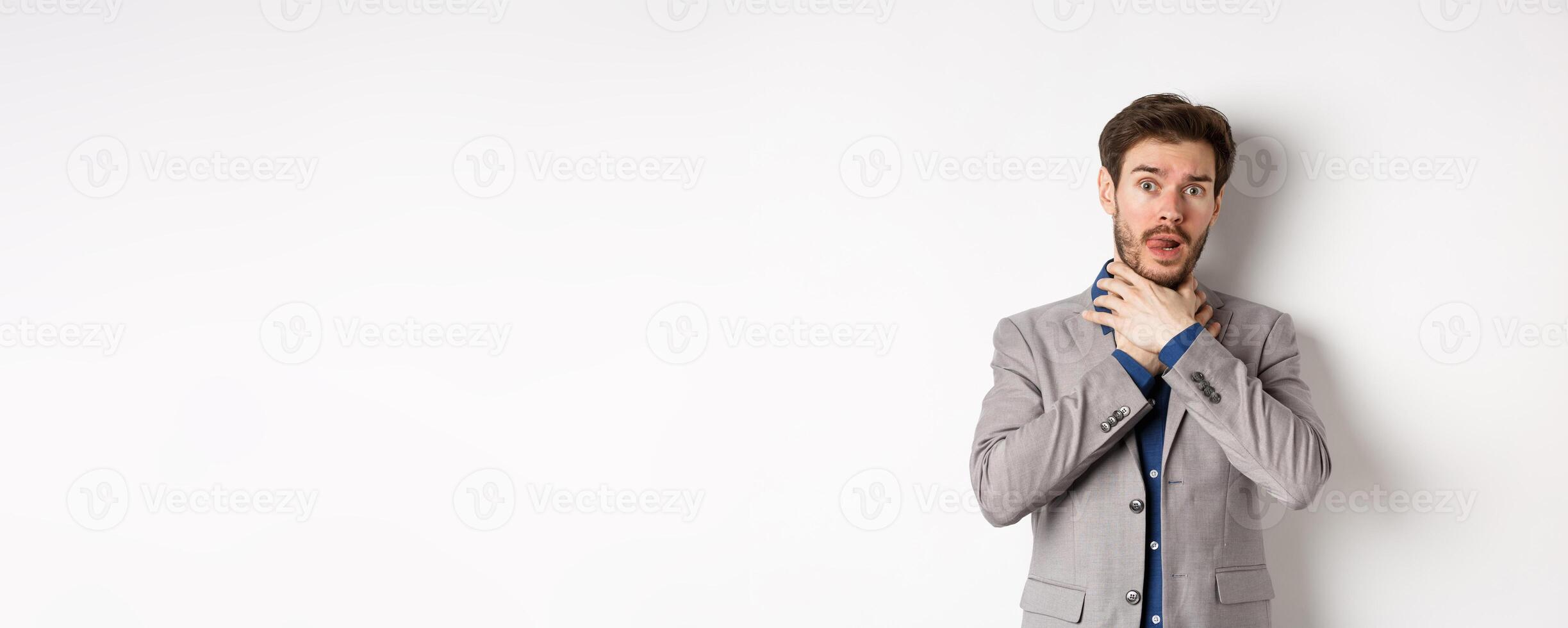 Distressed young man in business suit strangle himself and showing tongue, feeling sick with sore throat, standing on white background photo