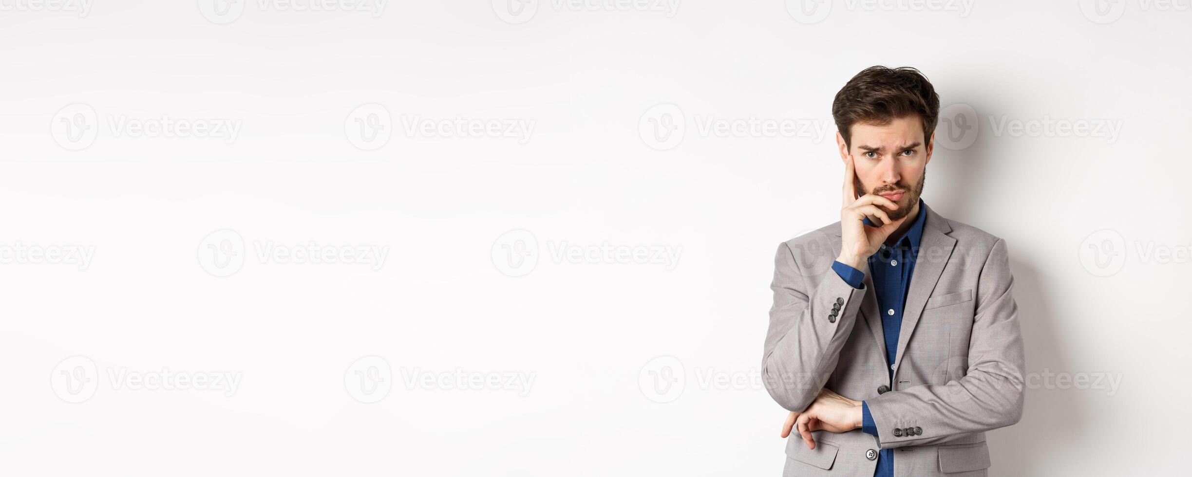 Troubled frowning business man in suit looking at camera pensive, thinking or making decision, standing on white background photo