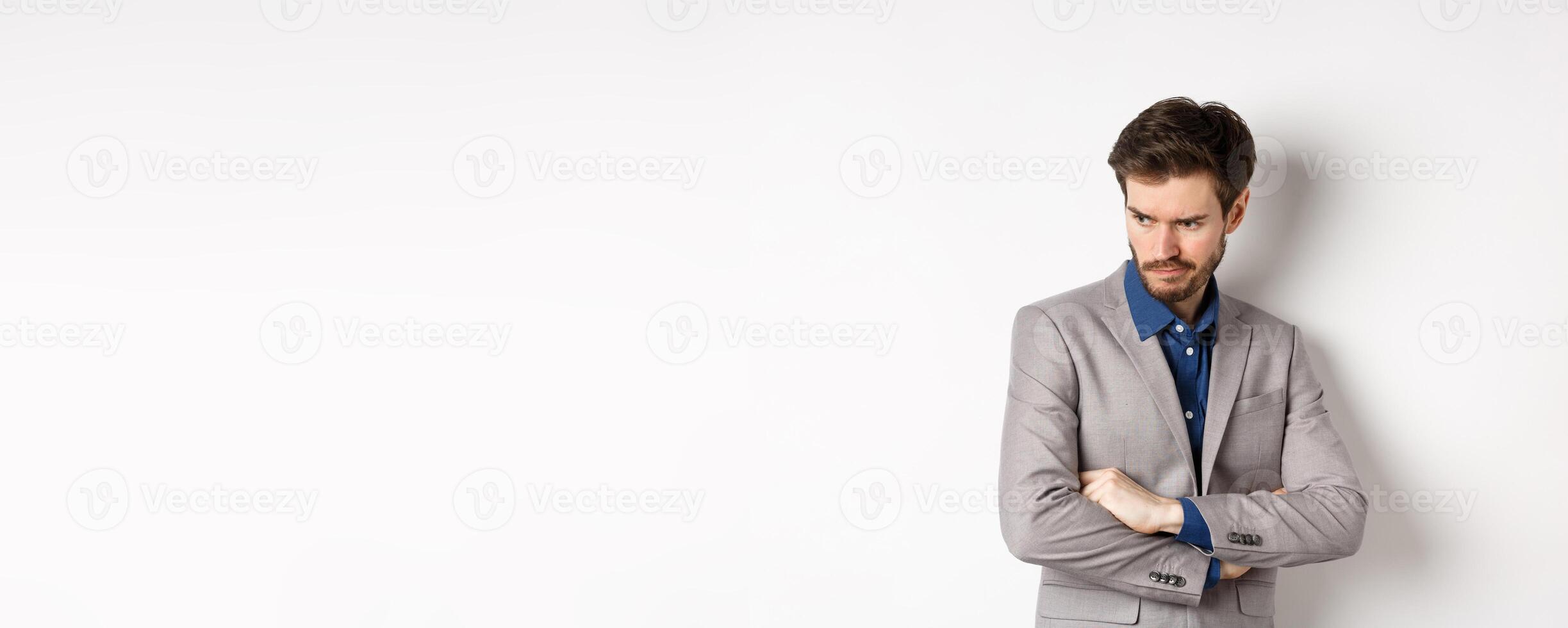Moody guy in suit looking aside with unfair face, cross arms on chest and frowning angry, standing upset against white background photo