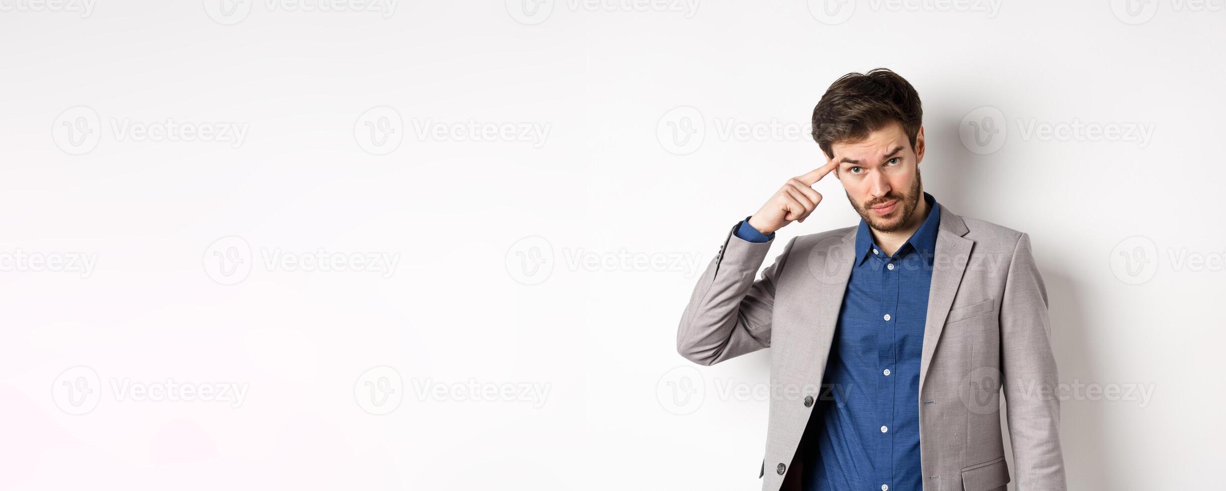 Annoyed male employer scolding personal, pointing finger at head, are you stupid out of your mind gesture, stare at camera from under forehead, standing in suit on white background photo