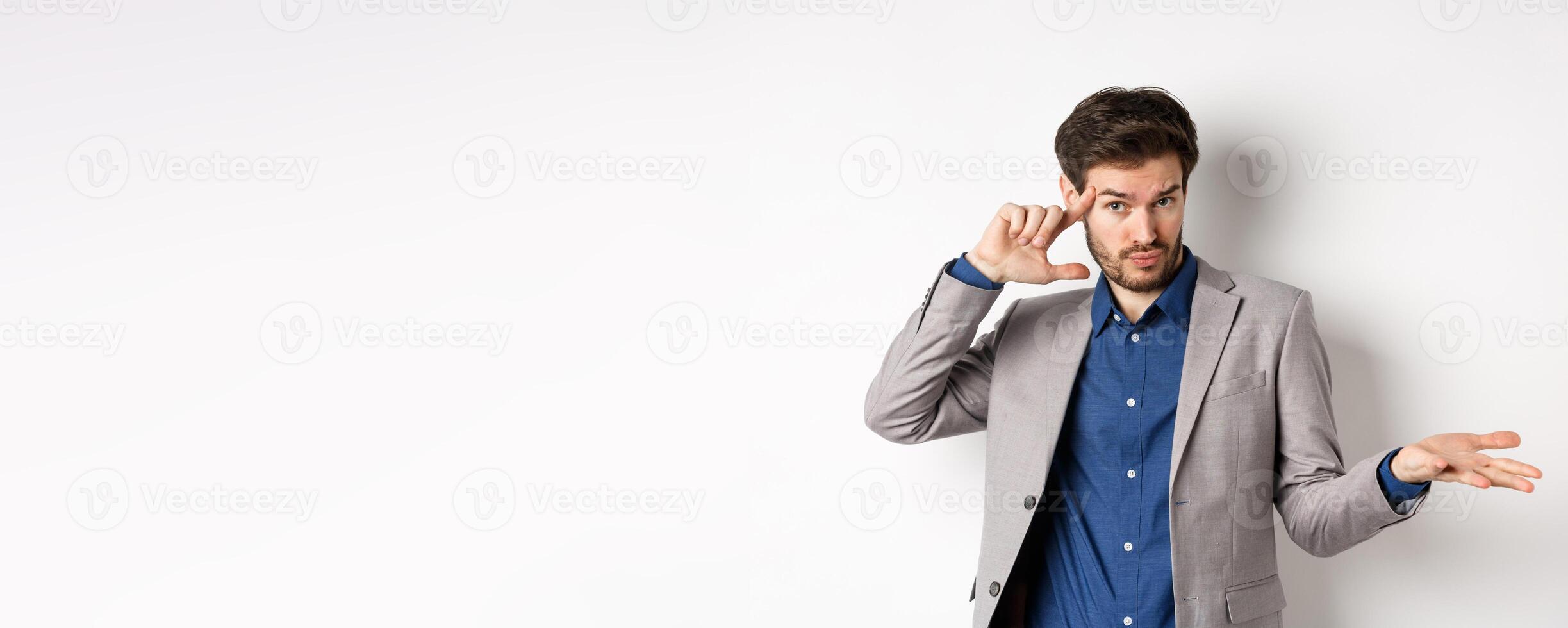 Are you crazy. Young man in suit pointing finger at head to scold strange or stupid mistake, shrugging shoulders confused, standing on white background photo