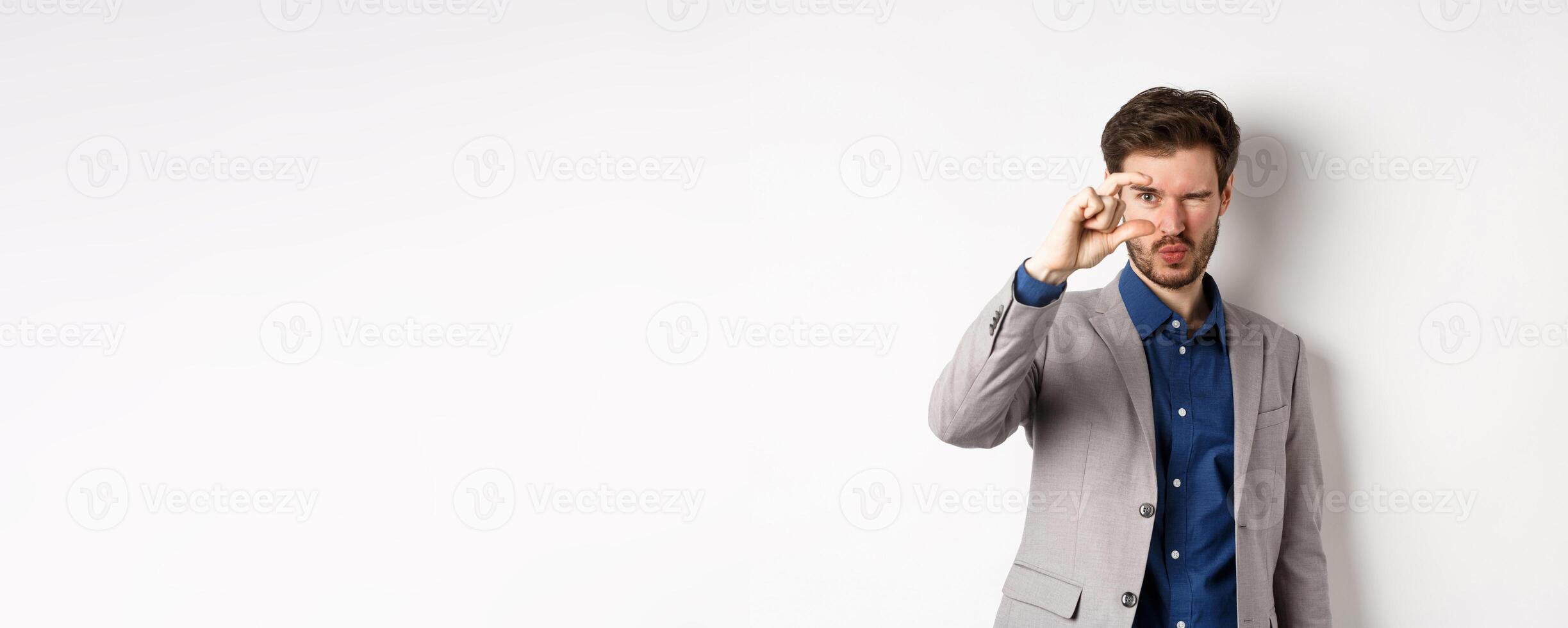Businessman trying to see something tiny, showing small thing size with fingers, standing in suit on white background photo