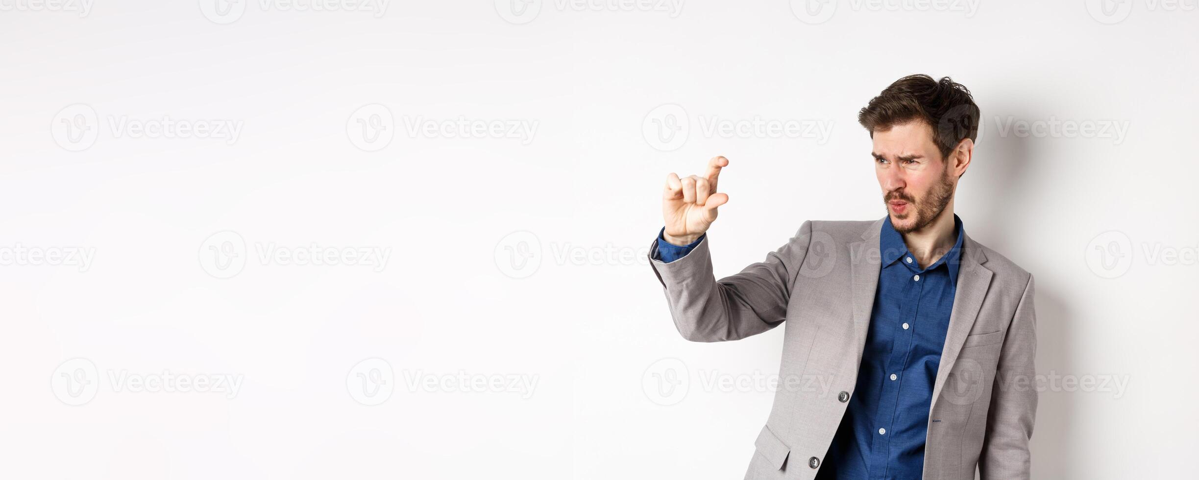 Businessman trying to see something small, showing little thing size with fingers, standing in suit on white background photo