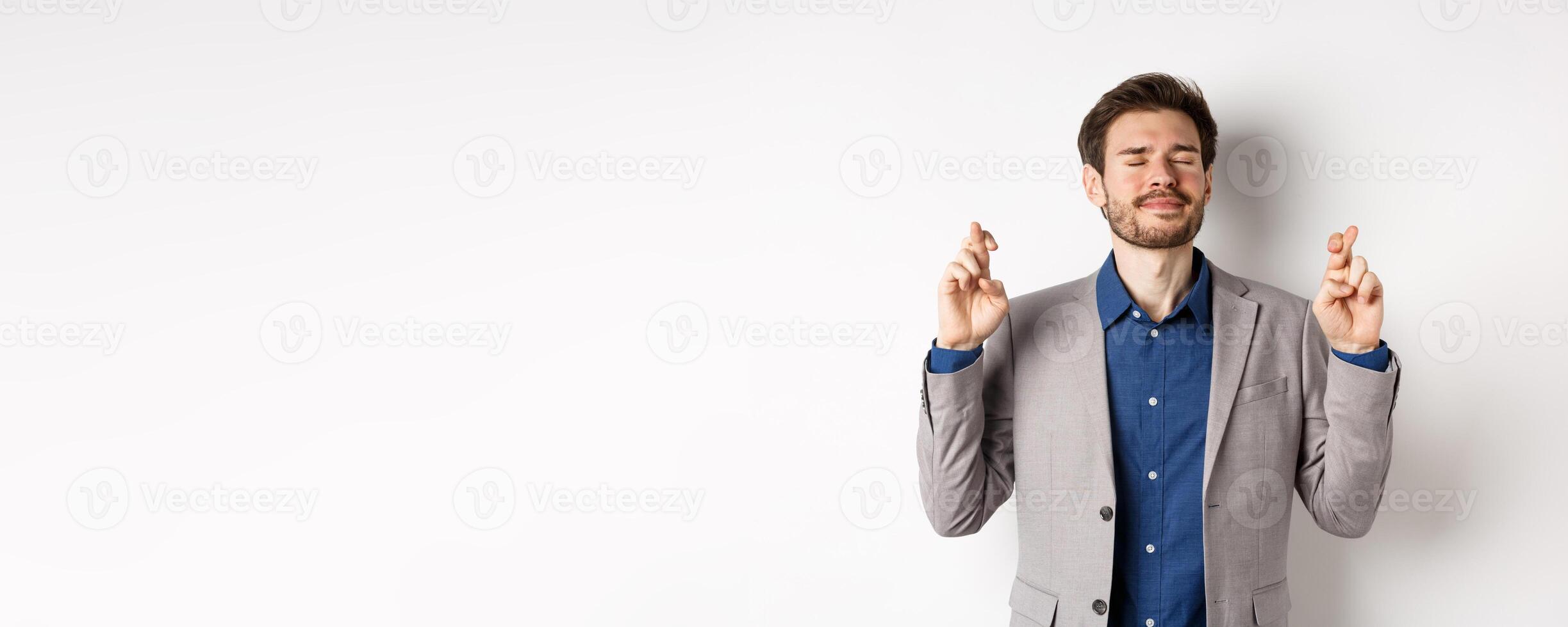 Nervous businessman in suit begging god, holding fingers crossed with eyes closed, pleading to win, making wish on white background photo