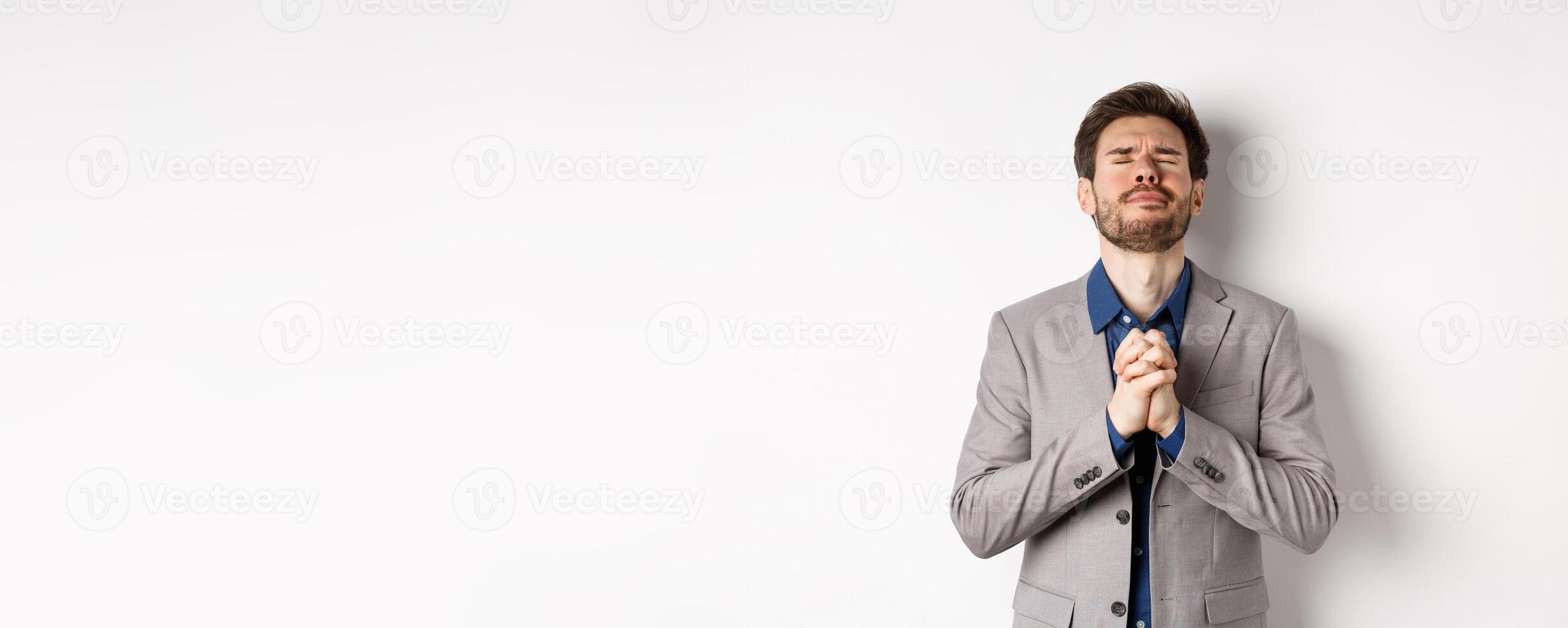 Nervous male entrepreneur in suit begging god, holding hands in pray with pleading expression, close eyes and make wish, asking for help, standing on white background photo