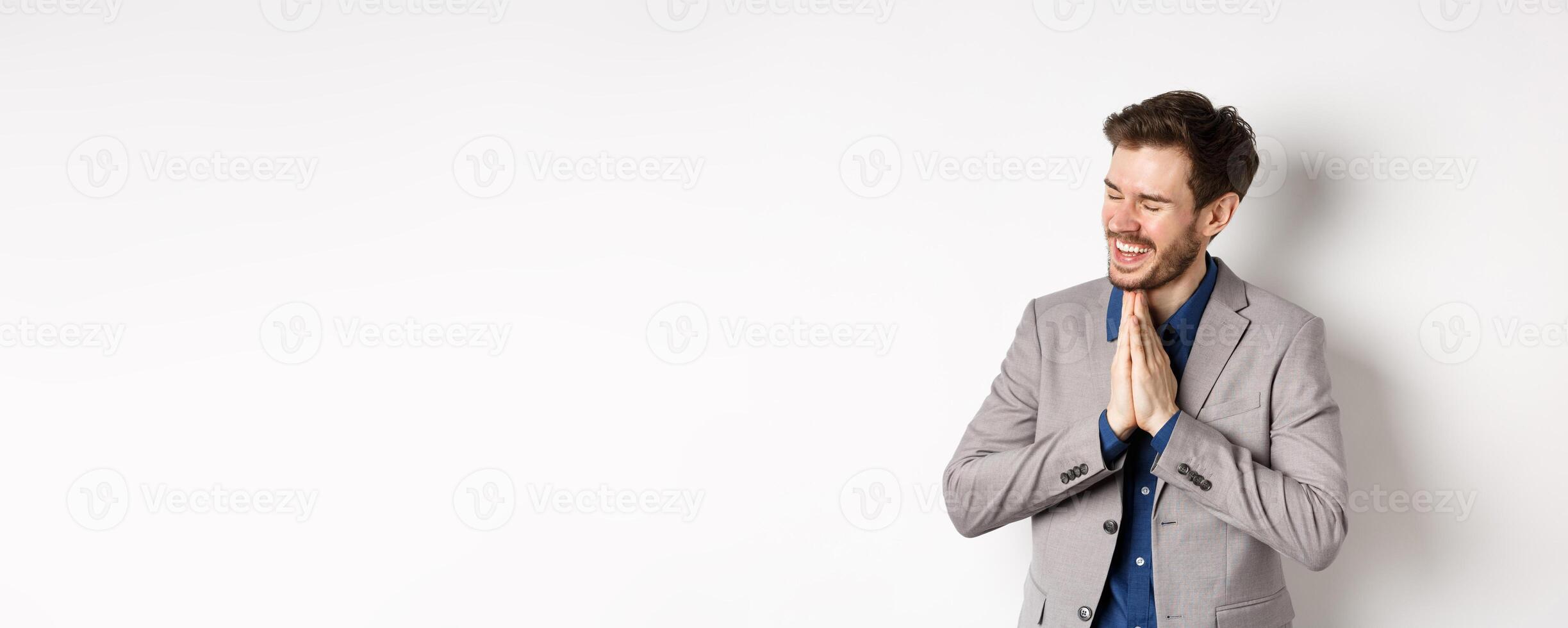 Happy and thankful businessman rejoicing, holding hands in begging or namaste sign, thanking for help, smiling relieved, standing in suit on white background photo