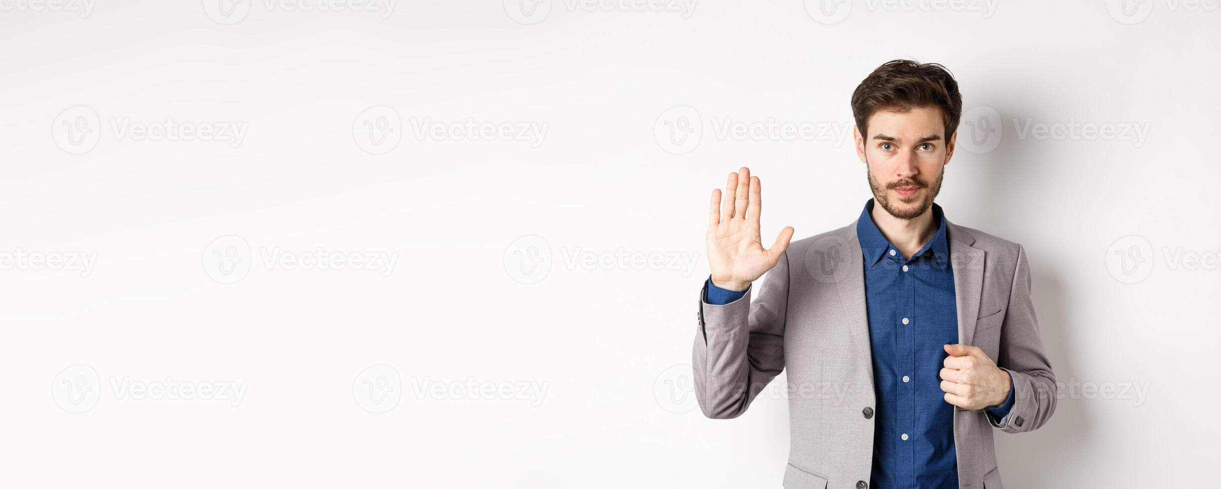 Businessman in formal suit raising hand up to say hello, contactless greeting, smiling friendly, standing on white background photo