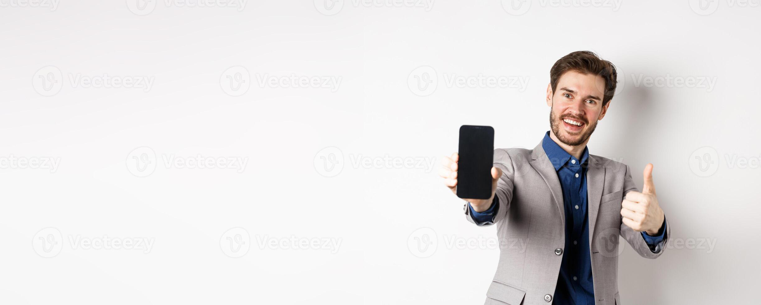 E-commerce and online shopping concept. Cheerful businessman in suit showing thumbs up and empty smartphone screen, recommending app, standing on white background photo