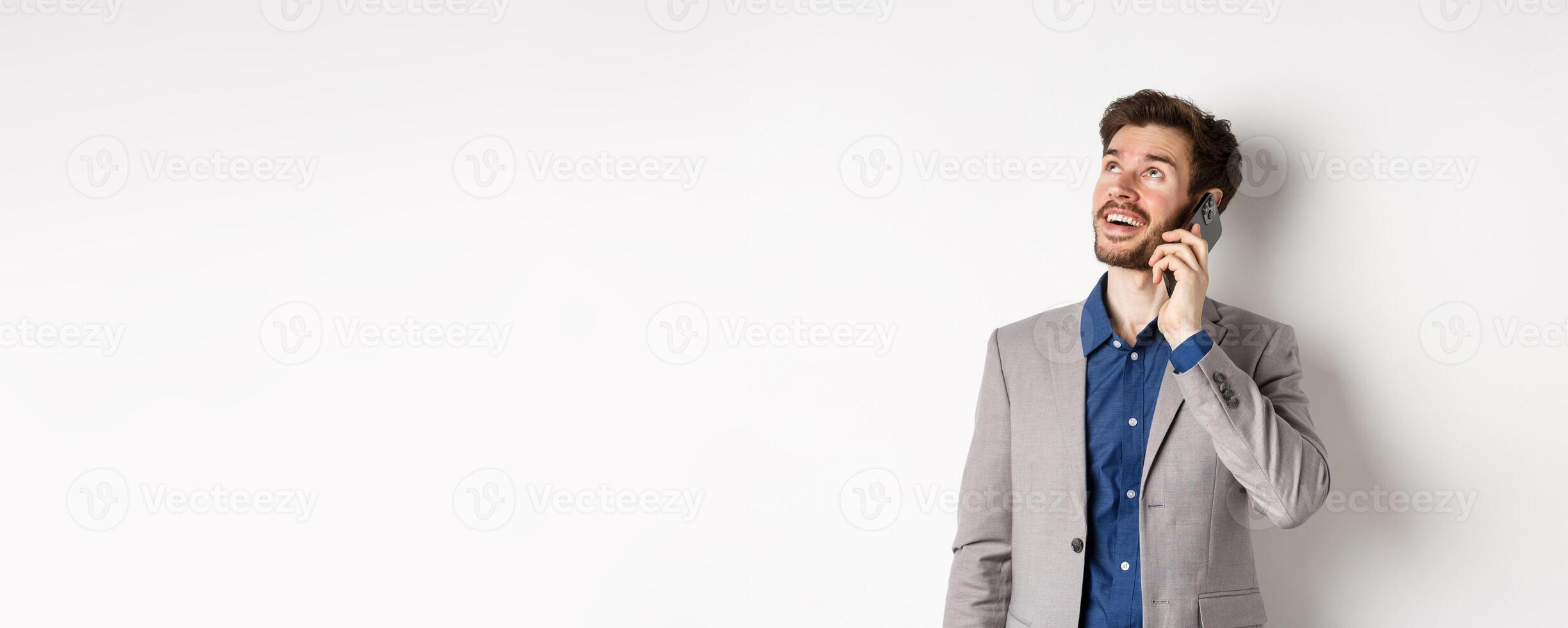 Carefree young man talking on mobile phone and smiling, looking up with dreamy face, standing in suit on white background photo