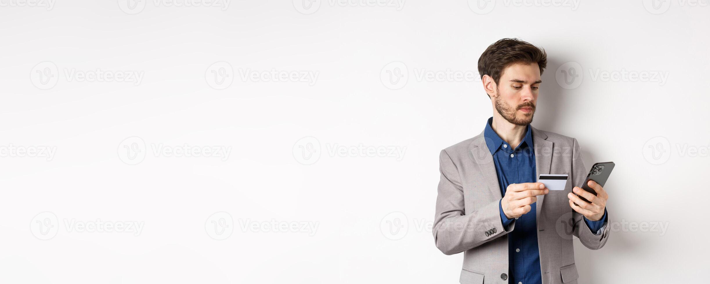 Online shopping. Serious man paying with credit card on smartphone, sending money, standing in suit on white background photo