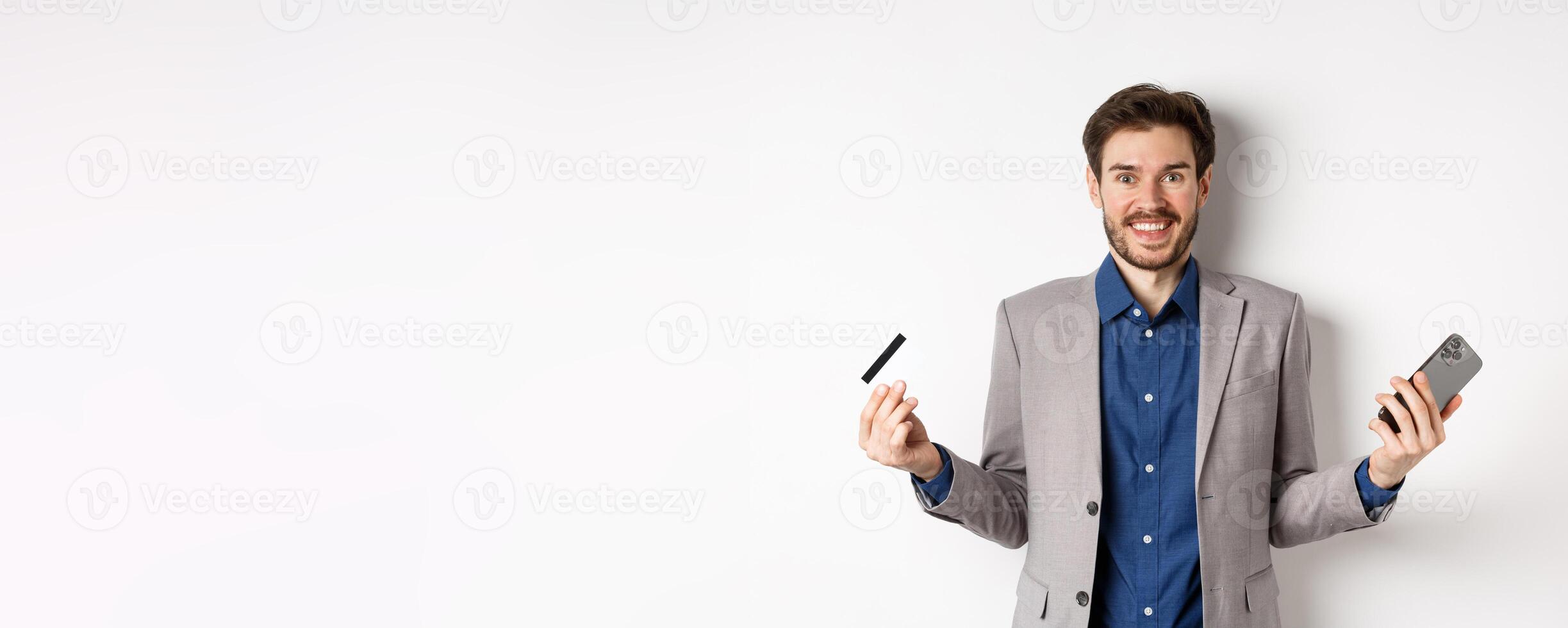 Online shopping. Excited man making money, smiling amazed, holding smartphone and credit card, standing against white background in suit photo