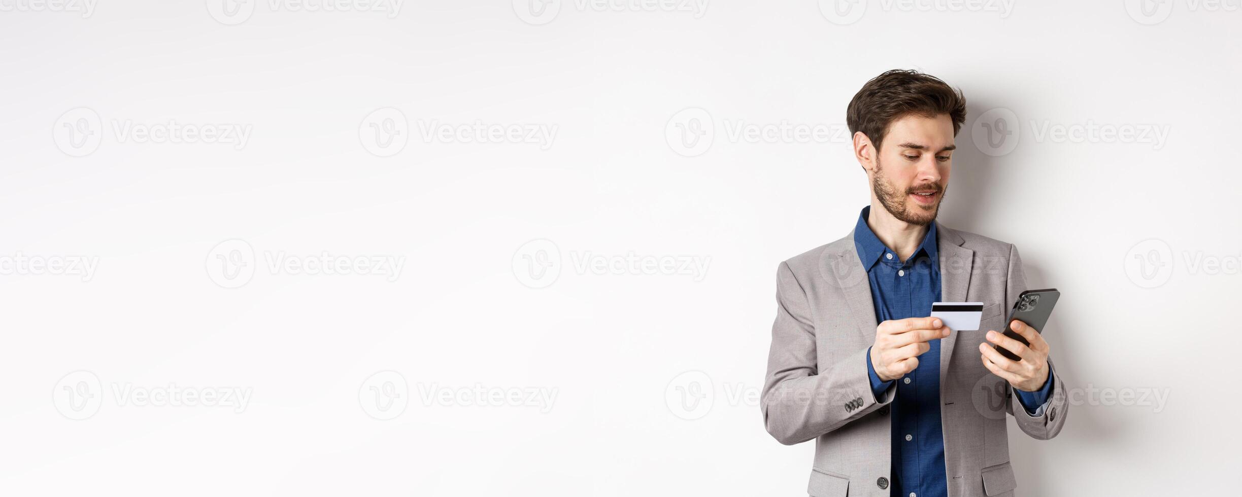 Online shopping. Smiling businessman paying with credit card on smartphone, sending money, standing in suit on white background photo