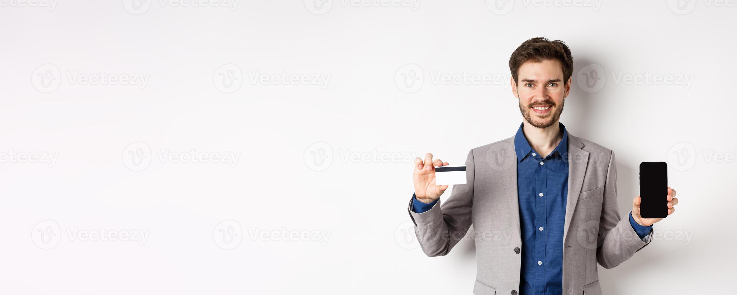 Online shopping. Smiling business man in suit showing plastic credit card with empty smartphone screen, standing against white background photo
