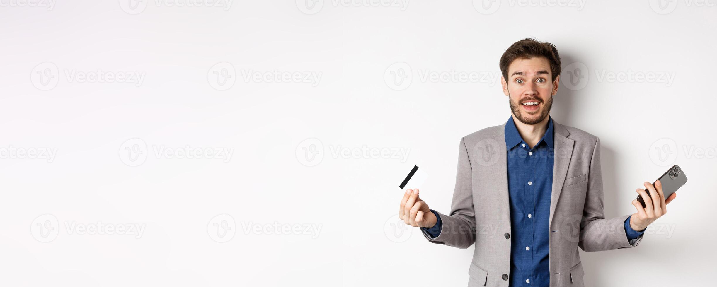 Online shopping. Surprised businessman holding plastic credit card and smartphone, making money in internet, standing in suit against white background photo